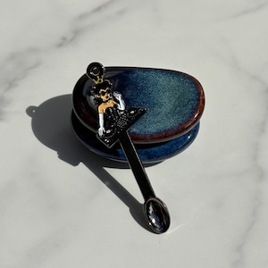 May include: A black metal spoon featuring a decorative enamel topper of a character wearing a crown, sunglasses, and white gloves while operating a DJ turntable. The spoon has a long, slender handle and a small, oval-shaped bowl at the end.