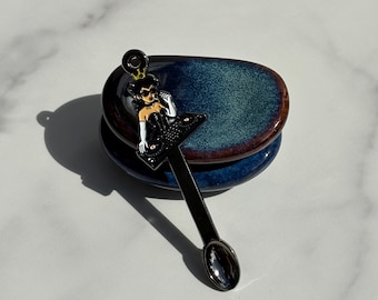 May include: A black metal spoon featuring a decorative enamel topper of a character wearing a crown, sunglasses, and white gloves while operating a DJ turntable. The spoon has a long, slender handle and a small, oval-shaped bowl at the end.