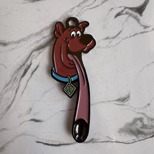 Puede incluir: Un llavero en forma de cabeza de Scooby-Doo de color granate con un collar azul y una etiqueta en forma de diamante con las letras "SD". El llavero tiene una lengua larga y rosa y un lazo marrón oscuro en la parte superior.