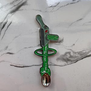 Puede incluir: Un pin de esmalte metálico que presenta un dispositivo de pistola de rayos gris y verde claro que emite un chorro de líquido verde brillante que fluye hacia abajo a través de un anillo verde hacia una pequeña cuchara plateada pulida.