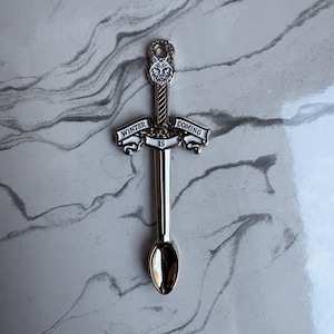 May include: A silver-toned metal spoon featuring a sword-shaped handle. The handle is topped with a wolf head emblem and includes a banner across the centre with the text "WINTER IS COMING" engraved in black lettering against a white background.