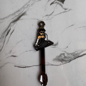 May include: A silver spoon with a decorative top. The top features a black and white illustration of a woman DJ wearing a black dress and sunglasses, standing behind a DJ mixing board. The spoon has a small, round bowl.