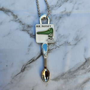 May include: A silver necklace with a charm that says "MR. NOSEY" and features a green cartoon character with a long nose. The charm is attached to a silver spoon.