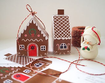 May include: Two gingerbread house shaped boxes with red and white trim, one with a candy cane design and the other with a wreath and candy dots. A small white snowman ornament with a red scarf and holly berries is in the foreground.