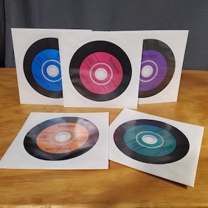 Könnte beinhalten: Eine Sammlung farbenfroher Verbatim Vinyl CDRs in weißen Papierhüllen. Die CDs haben einen schwarzen Außenring mit verschiedenfarbigen Zentren, darunter Blau, Pink, Lila, Orange und Türkis. Der Text "Verbatim Vinyl CDR" ist auf jeder Disc aufgedruckt.