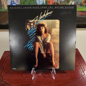 May include: A vinyl record album cover for the movie "Flashdance" featuring a woman in a gray sweater and red heels. The album cover is black with the movie title in a stylized font and blue and white graphics.