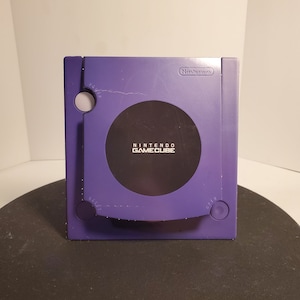 Peut inclure: Une console Nintendo GameCube violette avec un lecteur de disque noir et le texte "Nintendo GameCube" imprimé sur le devant.