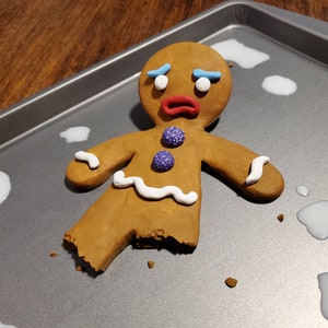 Könnte beinhalten: Ein Lebkuchenmann-Keks mit gebrochenem Bein liegt auf einem Backblech. Der Keks hat blaue Augenbrauen, weiße Zuckerguss-Akzente und lila Bonbonknöpfe. Der Mund ist rot, und er scheint traurig.