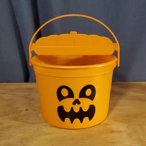 Puede incluir: Cubo de plástico naranja en forma de calabaza para pedir dulces con una cara de calabaza tallada negra y una tapa con bisagras.
