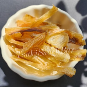 May include: A close-up of a small, cream-coloured, scalloped bowl filled with dried, golden-brown fish snacks. The snacks are thin and crispy. The text "Tsuki Snack Bar" is visible on the bowl. The bowl is on a dark surface.