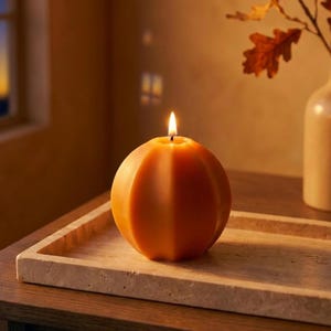 May include: A spherical, orange, pumpkin-shaped candle with vertical ridges and a lit wick, resting on a rectangular, light-colored, textured stone tray. The candle emits a warm glow, casting soft shadows across the surface of the tray.