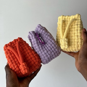May include: Three woven pouches in vibrant colors: orange, lavender, and yellow. Each pouch has a knotted handle and a textured, basket-weave design. The pouches are held up against a white background.