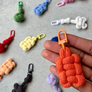 Może przedstawiać: Różnokolorowe, tkane breloczki do kluczy w różnych kolorach, w tym pomarańczowym, żółtym, zielonym, niebieskim, czerwonym i czarnym. Każdy breloczek posiada metalowy karabińczyk. Breloczki wykonane są z miękkiego, teksturowanego materiału.