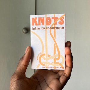 May include: A white booklet with the title "KNOTS intro to macrame" in orange text. The booklet features a diagram of a macrame knot in orange and white. The text "an instructional zine" is printed below the diagram.