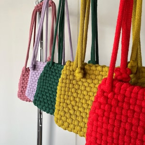 Op de afbeelding: Een rij kleurrijke, geweven touwtassen die aan een metalen stang hangen. De tassen zijn in tinten roze, lavendel, groen, geel en rood. Elke tas heeft een bijpassende touwhandgreep. De tassen zijn gemaakt van een dik, getextureerd materiaal.