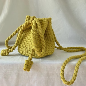 May include: A yellow, hand-knitted bucket bag with a braided rope strap and drawstring closure. The bag is made of a textured, woven material and features a small, matching coin purse. The bag's strap is approximately 40 inches long.