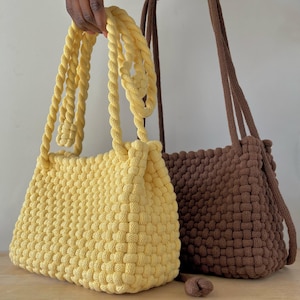 May include: Two handmade macrame handbags. One is a light yellow color, and the other is brown. Both bags have woven bodies and thick, twisted rope handles. The yellow bag is held up by a person's hand.
