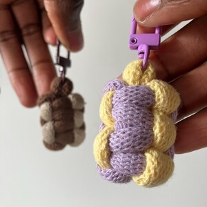 May include: Two crocheted keychains with purple and yellow yarn. The keychain on the left is brown and beige. Both keychains have a purple metal clasp.