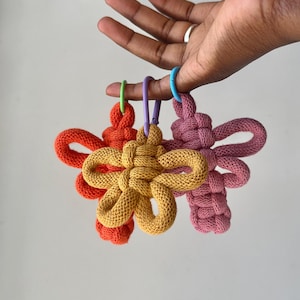 以下が含まれることがあります： オレンジ、マスタードイエロー、ダスティローズの織りコードで作られた3つの花型キーホルダー。各キーホルダーには、結び目のある布製の花のデザインの中心に、緑、紫、または青の小さなプラスチック製カラビナが取り付けられています。