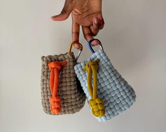 May include: Two small rectangular pouches made of woven cord. One pouch is tan with an orange drawstring and an orange carabiner. The other pouch is light blue with a mustard yellow drawstring and a blue carabiner. Both feature a knotted cord.