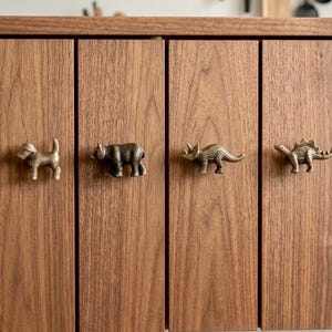 Può includere: Quattro pomelli per armadietti in metallo color ottone a forma di animali, tra cui un cane, un orso, un triceratopo e uno stegosauro, montati su quattro ante di armadietti in legno verticale con venature visibili e finitura marrone.