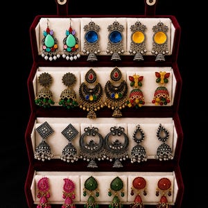 May include: A velvet jewelry display case containing four rows of various dangling earrings. The collection features diverse designs including teardrop shapes, circular studs, and bell-shaped jhumka styles in vibrant colors like green, blue, yellow, red, and pink.