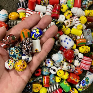Może przedstawiać: Duża kolekcja różnorodnych szklanych koralików o różnych kształtach, rozmiarach i kolorach, w tym czerwonym, żółtym, niebieskim, białym, zielonym i czarnym, z rozmaitymi wzorami, takimi jak paski, motywy kwiatowe, kropki i marmurkowe wykończenia.
