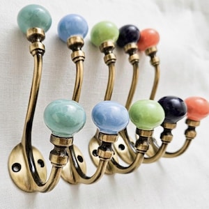 Op de afbeelding: Een set van vijf wandhaken met gebogen armen van antiek messing en ronde keramische knoppen in diverse kleuren, waaronder mintgroen, lichtblauw, limoengroen, donker marineblauw en feloranje, gerangschikt in een verspringende rij.