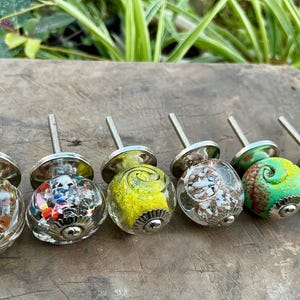 Puede incluir: Una fila de tiradores decorativos de cristal con herrajes plateados. Los tiradores presentan diseños coloridos, incluyendo espirales y patrones abstractos. Cada tirador tiene aproximadamente 4 cm de diámetro, añadiendo un toque único a los muebles.