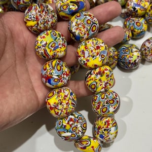 Könnte beinhalten: Eine Sammlung runder Glasperlen mit einem lebendigen Millefiori-Muster auf leuchtend gelbem Grund. Die Perlen sind mit komplexen, mehrfarbigen, blumenartigen Formen in Rot-, Blau-, Weiß- und Schwarztönen verziert, was jeder einzelnen Perle ein dichtes, mosaikartiges Aussehen verleiht.