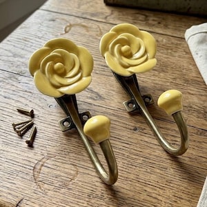 May include: Two wall hooks featuring pale yellow ceramic rose-shaped finials and matching yellow ball-tipped ends on curved antique brass-toned metal arms. Each hook includes a mounting plate with two screw holes, accompanied by four small metal screws.
