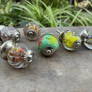 Puede incluir: Una colección de tiradores decorativos para armarios con una variedad de diseños coloridos. Los tiradores presentan globos de vidrio transparente con patrones internos de rojo, amarillo y azul, y algunos tienen una superficie verde o estampada. Cada tirador tiene una base y un tornillo de metal plateado.