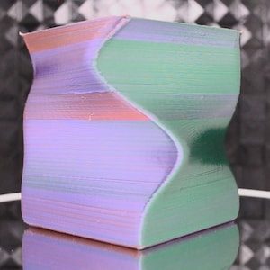 May include: A 3D printed vase with a wavy, abstract design. The vase is made of plastic and is painted in shades of green, purple, and pink.