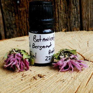 May include: A small, dark brown glass bottle with a black cap. The bottle is labeled "Botanicals Bergamot Bath 5ml 2215". The bottle is sitting on a wooden surface with two purple flowers.