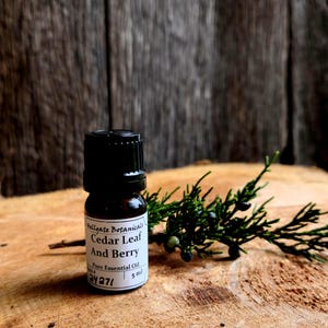 May include: A small brown glass bottle with a black cap, labeled "Cedar Leaf And Berry Pure Essential Oil 5 ml." The bottle sits on a wooden surface next to a sprig of green foliage with small blue berries. The background is a wooden wall.