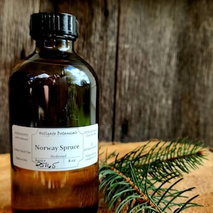 May include: A brown glass bottle with a black cap, labelled "Hellgate Botanicals Norway Spruce Hydrosol 8 oz." A sprig of green spruce needles rests beside the bottle on a wooden surface. The background is a wooden wall.