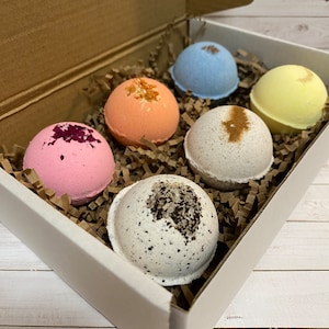 May include: A white box filled with six colorful bath bombs. The bath bombs are various colors, including pink, orange, blue, white, and yellow. Some are decorated with dried flowers or spices. The box is lined with brown crinkle paper.