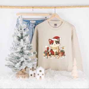 May include: A beige sweatshirt featuring a Christmas cat illustration wearing a Santa hat and scarf, surrounded by wrapped gifts and string lights. The sweatshirt is hanging on a wooden hanger with a pair of blue jeans.