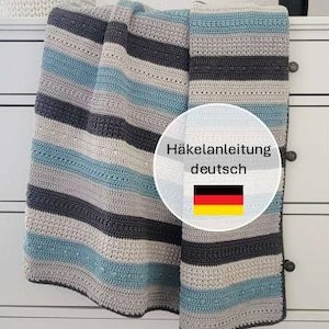 Könnte beinhalten: Eine handgefertigte Häkeldecke mit horizontalen Streifen in Grau-, Blau- und Cremetönen. Die Decke ist über einer weißen Kommode drapiert. Das Bild zeigt auch einen kleinen Korb und eine Vase mit rosa Blumen.