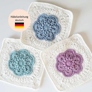 以下が含まれることがあります： 花柄のデザインが施された、正方形の編み物コースター3枚。各コースターは白い縁取りと、青、ティール、紫色の中心の花柄が特徴です。画像にはドイツ語のテキストが含まれています。