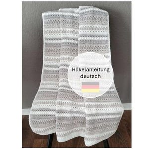 May include: A folded, handmade crochet blanket in shades of gray and white. The blanket features a striped pattern and is draped over a dark surface. A circular graphic with German text and flag is visible.