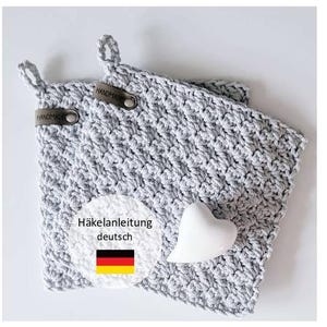 Könnte beinhalten: Zwei gehäkelte Topflappen in Grau mit Schlaufengriffen und Lederetiketten mit der Aufschrift "HANDMADE". Eine herzförmige Dekoration in Weiß und eine Grafik mit deutschem Text und Flagge sind ebenfalls vorhanden.