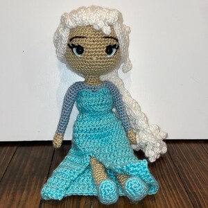 May include: Handmade crocheted doll with a light brown face, blue eyes, and white hair in a braid. The doll is dressed in a blue dress with long sleeves. The doll is sitting on a wooden surface.