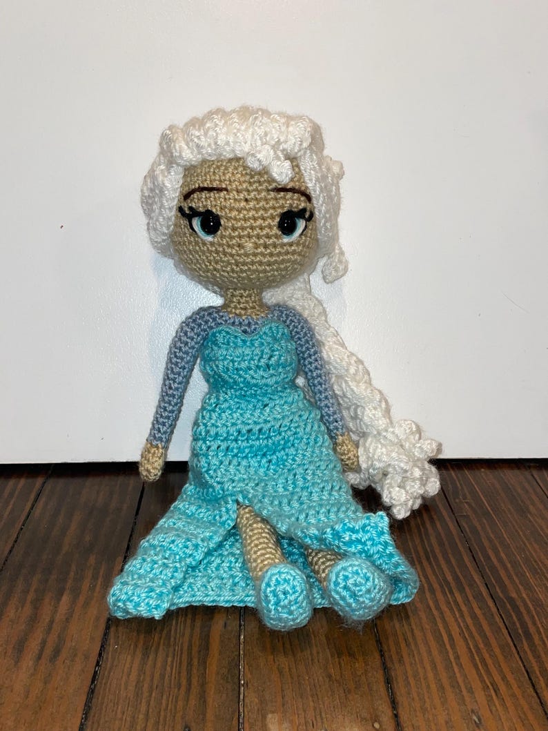 May include: Handmade crocheted doll with a light brown face, blue eyes, and white hair in a braid. The doll is dressed in a blue dress with long sleeves. The doll is sitting on a wooden surface.