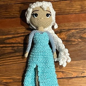 May include: A handmade crochet doll with a light skin tone face, black eyes, and white hair. The doll is dressed in a light blue dress with a high slit. The doll is approximately 25 cm tall.