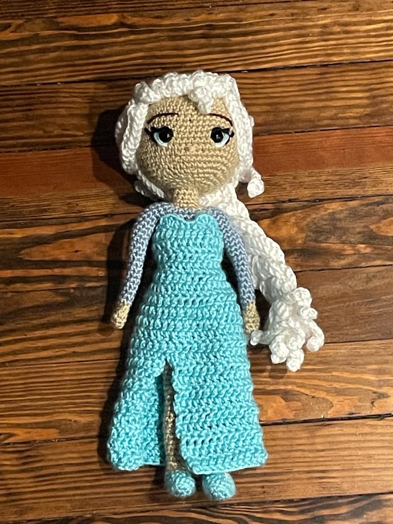 May include: A handmade crochet doll with a light skin tone face, black eyes, and white hair. The doll is dressed in a light blue dress with a high slit. The doll is approximately 25 cm tall.