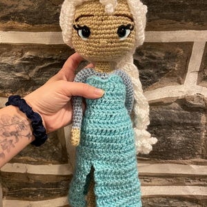 May include: A handmade crochet doll with a light tan face, large black eyes, and white hair styled in a braid. The doll wears a light blue dress with a slit. The doll is being held by a person.