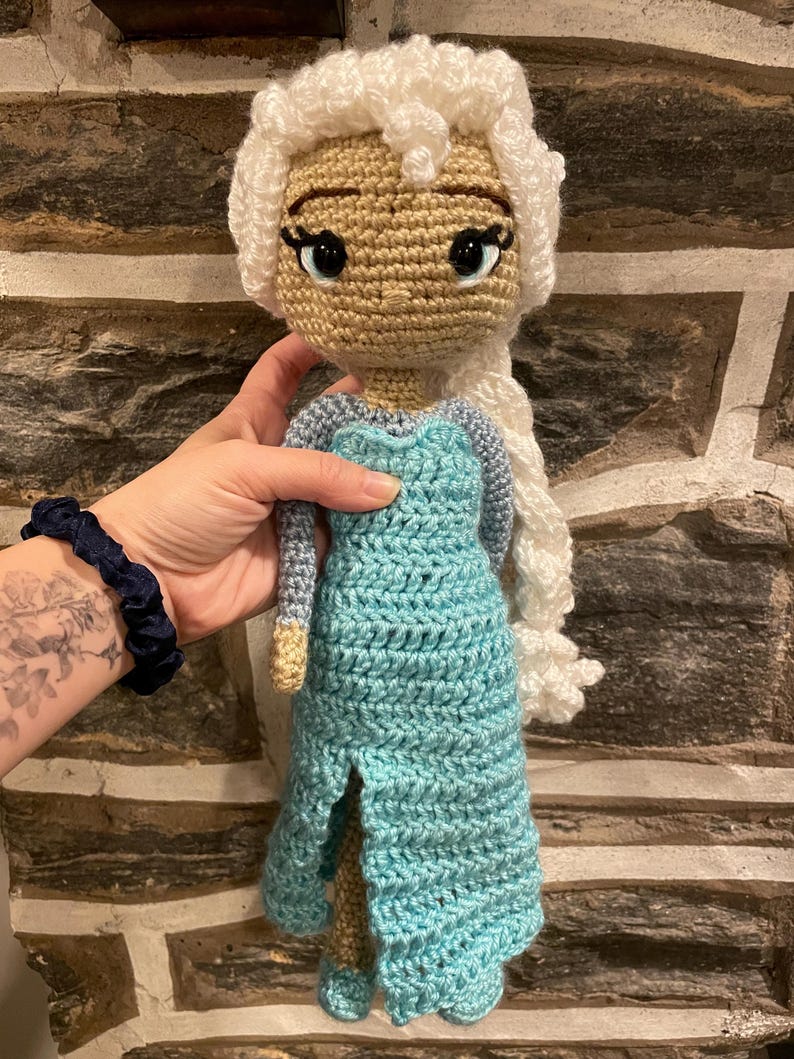 May include: A handmade crochet doll with a light tan face, large black eyes, and white hair styled in a braid. The doll wears a light blue dress with a slit. The doll is being held by a person.
