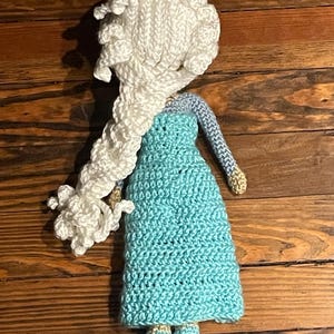 May include: A handmade crochet doll with a white braided hairstyle and a turquoise dress. The doll has light blue sleeves and gold accents on the shoes. The doll is placed on a wooden surface.