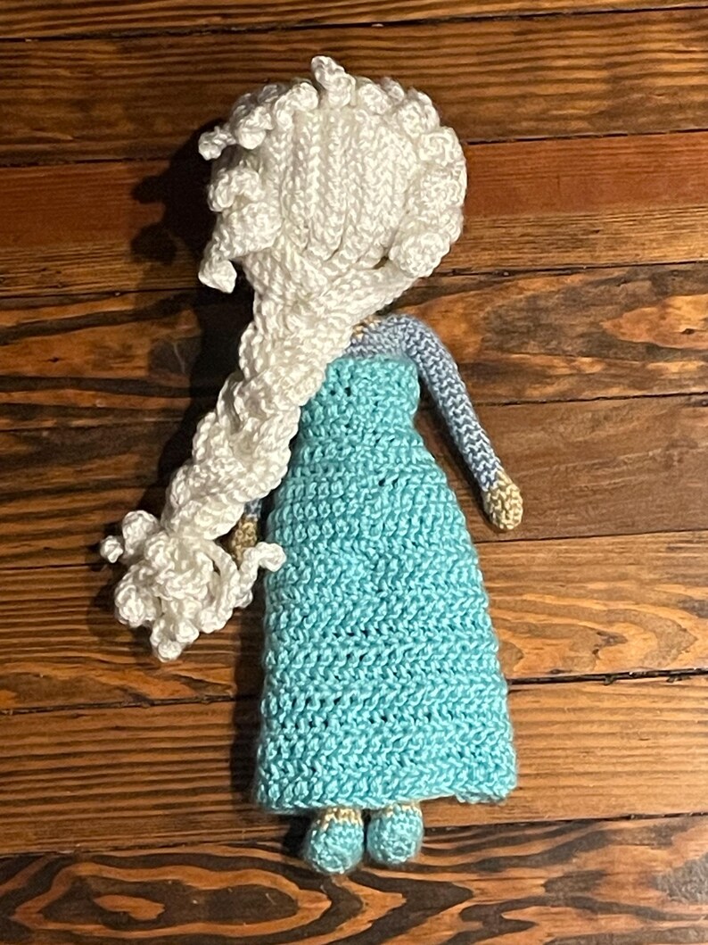 May include: A handmade crochet doll with a white braided hairstyle and a turquoise dress. The doll has light blue sleeves and gold accents on the shoes. The doll is placed on a wooden surface.